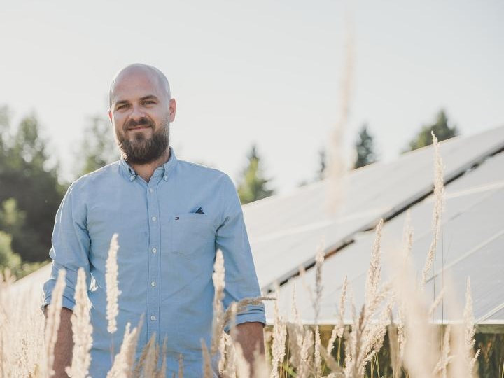 Ein junger Mann grinst in die Kamera. Hinter ihm ist eine Solaranlage zu sehen