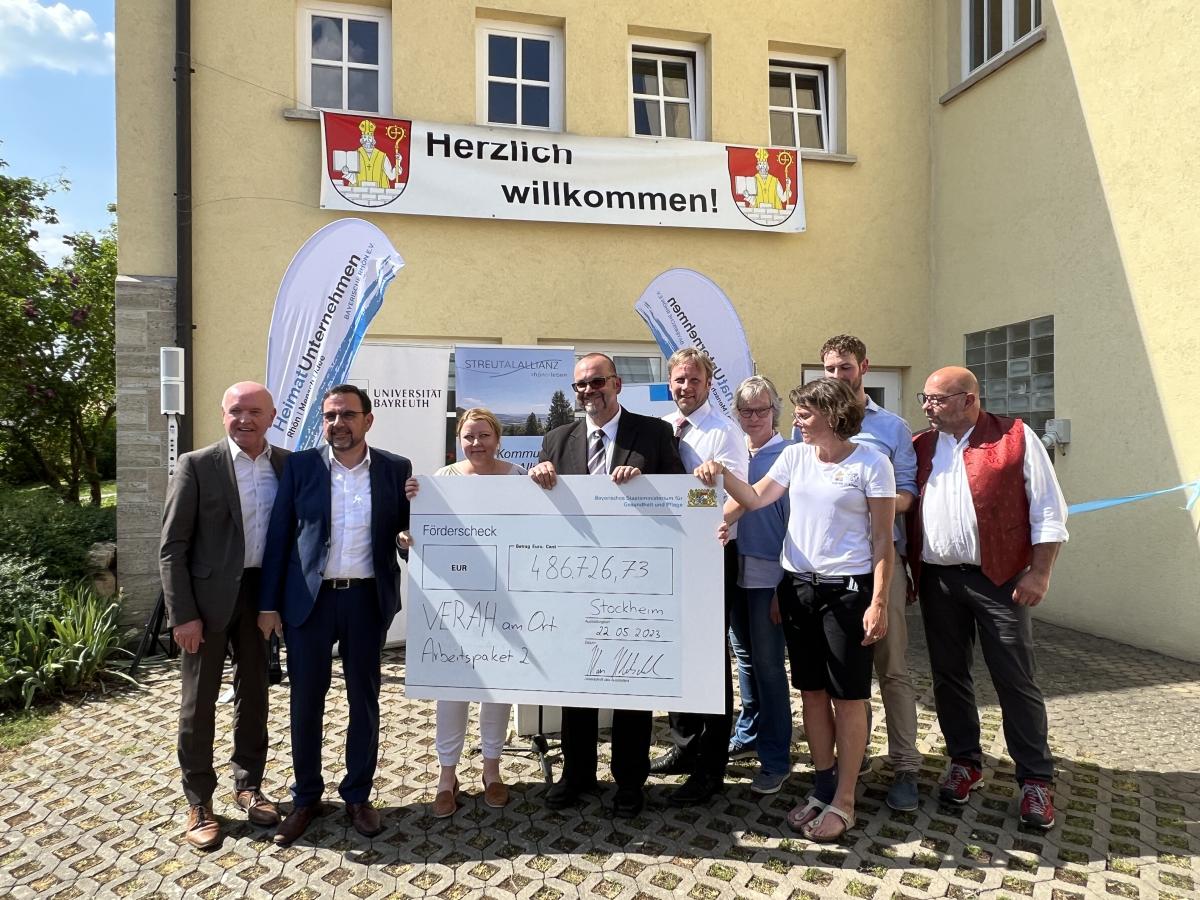 Ein übergroßer Scheck wird symbolisch übergeben. Eine Gruppe steht vor einem Haus