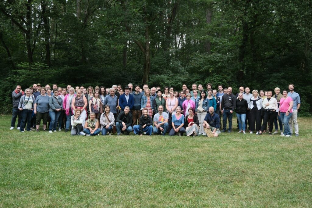 Sehr viele bunt gekleidete Menschen stehen zusammen auf eienr Wiese und strahlen.