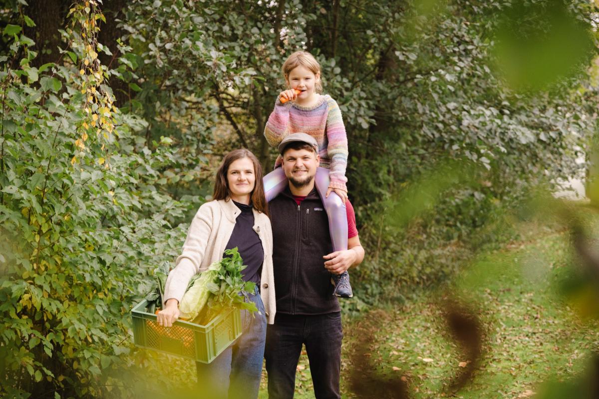 Bild einer Familie: Die Frau steht mit einer Gemüsekiste da, der Mann trägt ein Bild auf den Schultern