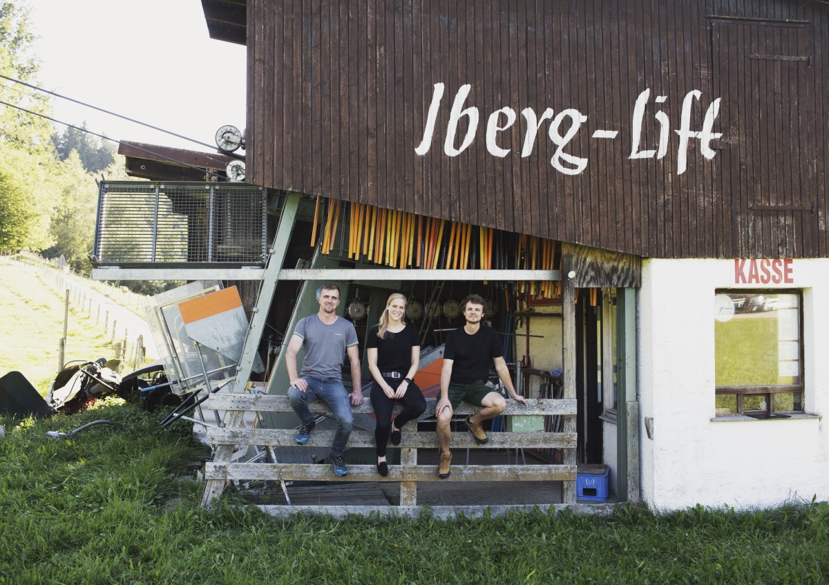 Drei junge Menschen sitzen auf einem Holzzaun vor dem Iberg-Lift Maierhöfen/Riedholz