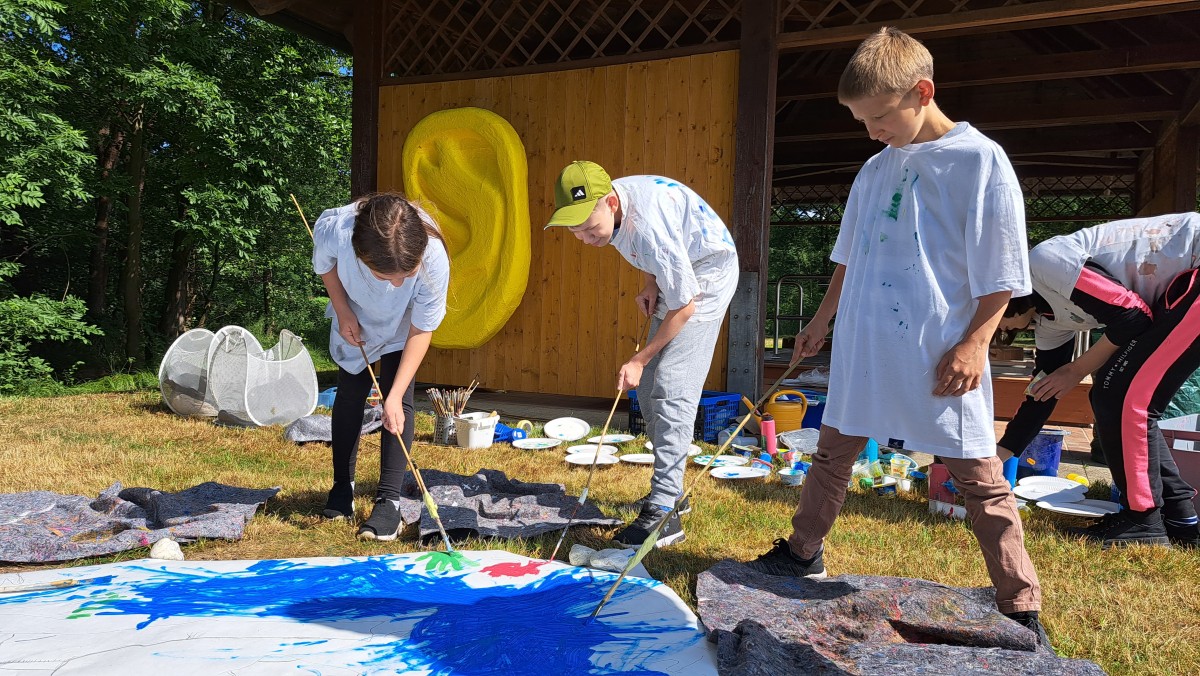 Drei Kinder in großen weißen T-Shirts malen zusammen an einem Bild. Das Bild liegt auf dem Rasen und die Kinder benutzen extralange Pinsel.