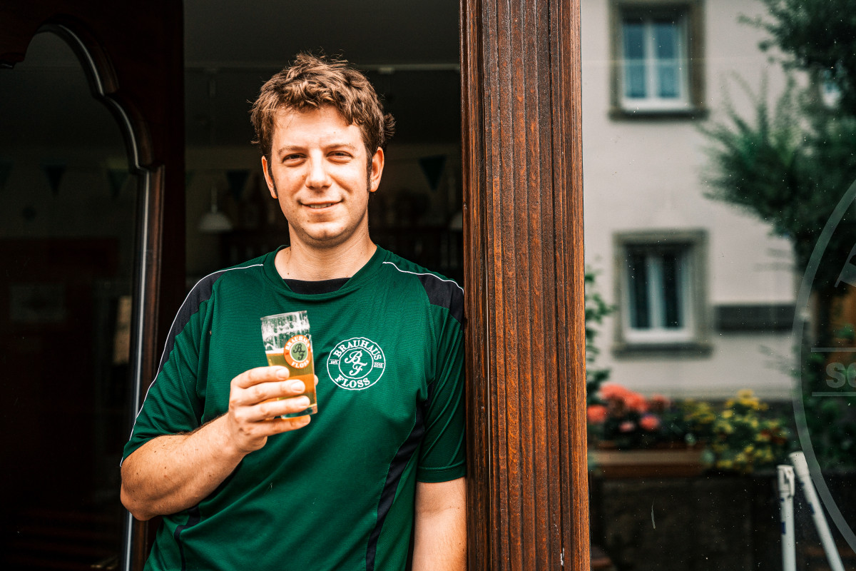 Ein Mann im grünen Shirt steht vor einem großen Fenster. Er hält ein Bier in der Hand und lächelt in die Kamera.