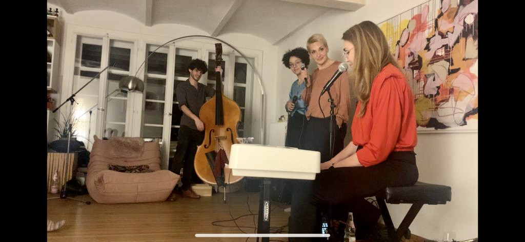 Ein Wohnzimmer. Ein Mann am Kontrabass stehend, eine Frau am Piano sitzend, zwei Frauen am Mikrofon stehend