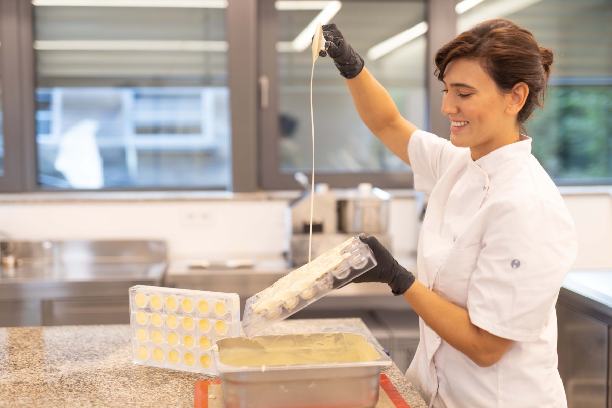 Patricia steht mit zusammengebundenen Haaren und in Kochkleidung in ihrer Manufaktur. Mit einem Lächeln auf den Lippen befüllt sie eine Form für Pralinen mit weißer Schokolade.