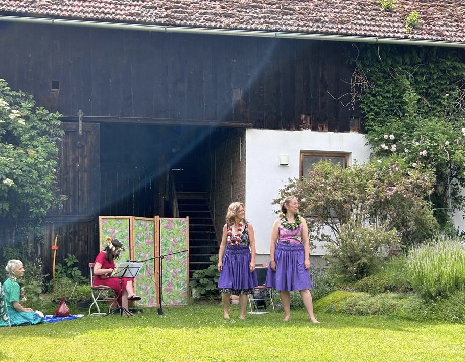 Zwei Frauen tanzen mit Blumenkleidern vor einem sehr großen alten Holzhaus