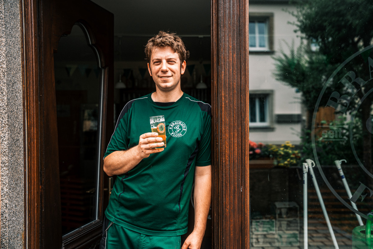 Ein Mann im grünen Shirt steht vor einem großen Fenster. Er hält ein Bier in der Hand und lächelt freundlich in die Kamera.