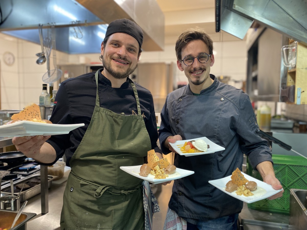 Zwei Männer in einer Küche haben jeweils 2 angerichtete Speisen-Teller in der Hand